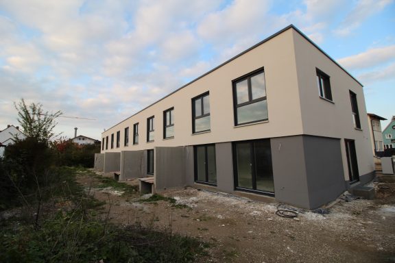 Moderne Gebäude mit mehreren Fenstern und grauer Fassade, umgeben von unbefestigtem Gelände.