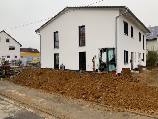Neues, zweigeschossiges, weißes Haus mit Erdhaufen und Baumaschinen davor.