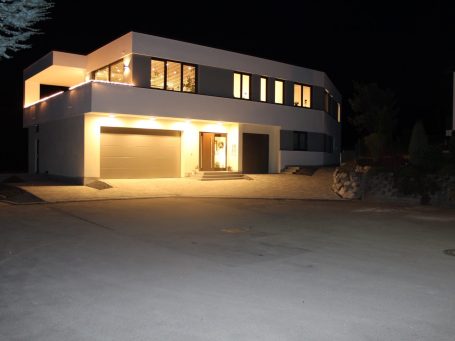 Modernes Haus mit Beleuchtung, in der Nacht, umgeben von einer Auffahrt und Bäumen.